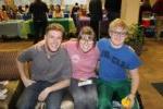 three smiling students sitting on a couch with information booths in the background